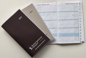 Two closed annual planners (burgundy and gray) labeled 2027 and 2021 beside an open weekly calendar showing December 2026 and January 2027 on a light surface of a desk; includes a bank logo on the burgundy cover (Bezirks-Sparkasse Dielsdorf).